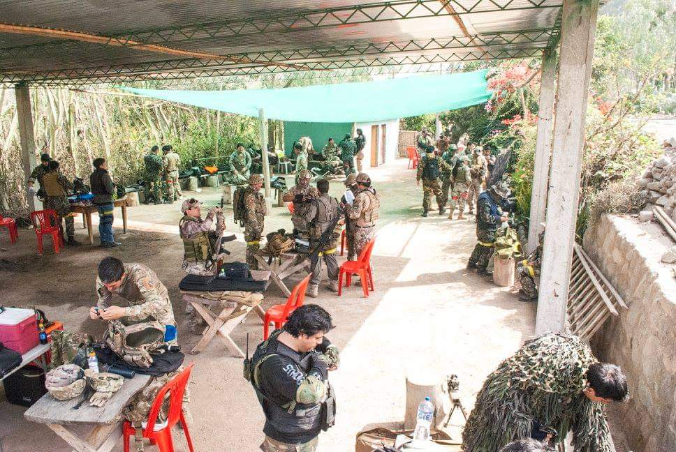 Jugadores de airsoft con uniformes militares preparan su equipo bajo un gran cobertizo exterior.