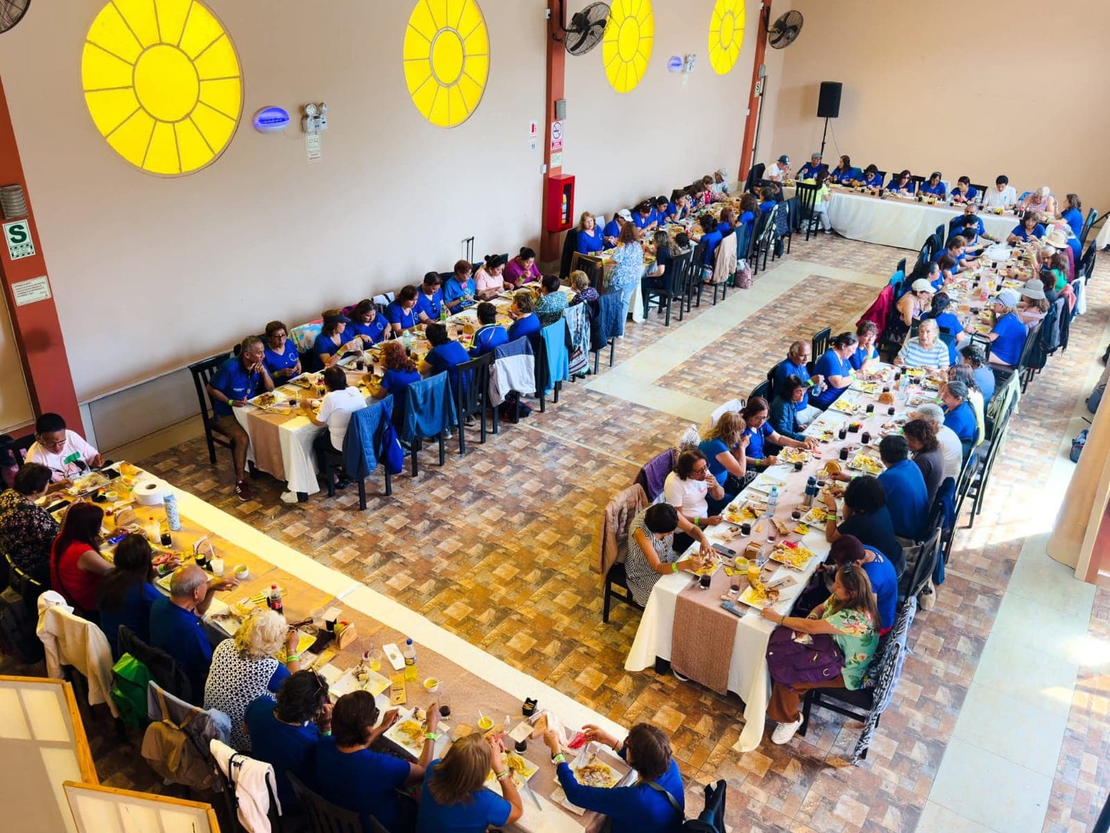 Personas almorzando en mesas largas dentro de un gran salón con ventanas circulares amarillas.