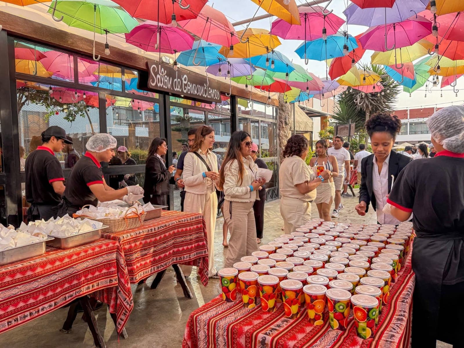 Personal sirviendo comida y jugos de frutas bajo un techo decorado con paraguas coloridos.