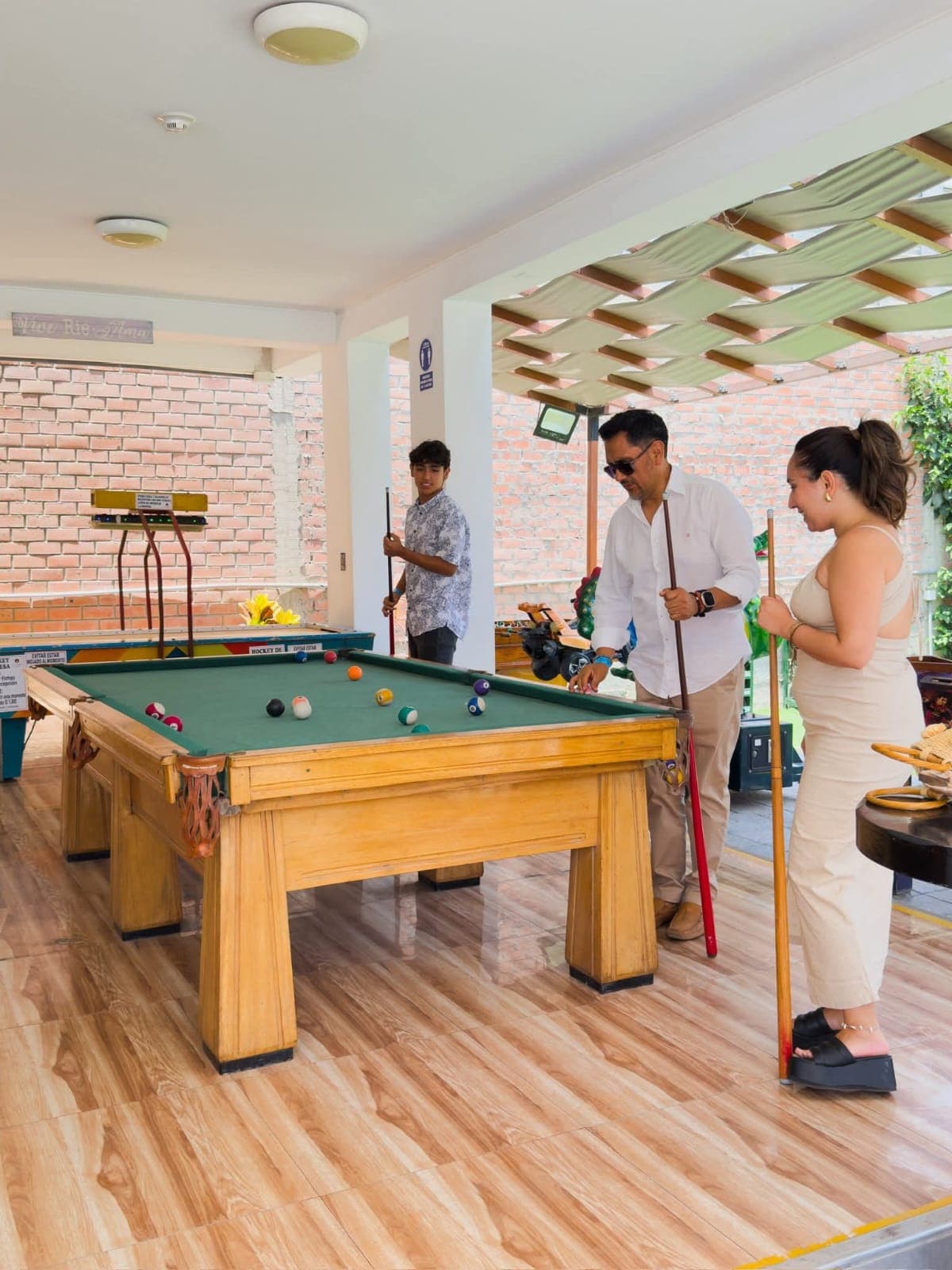 Tres personas juegan al billar en una mesa de madera en un patio exterior techado.