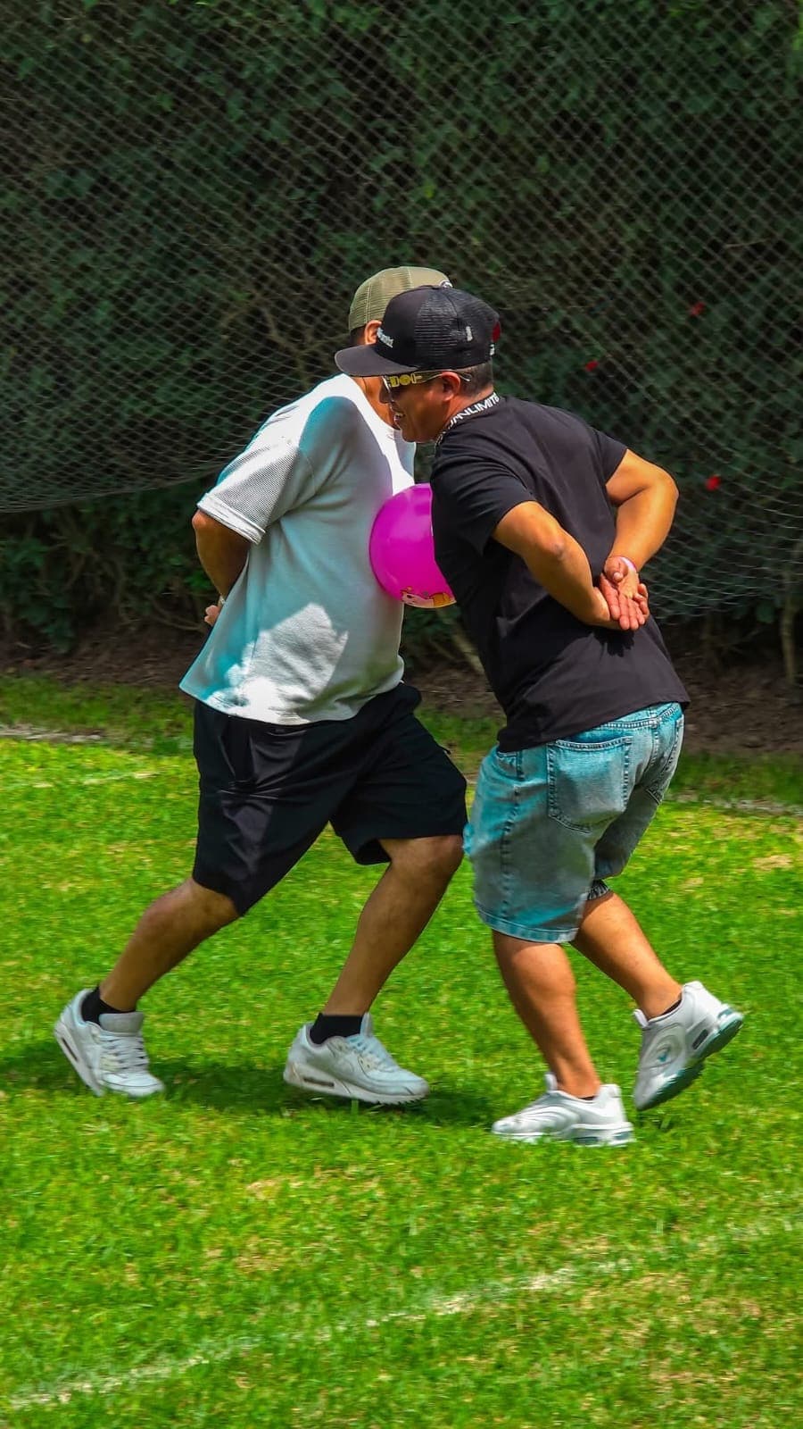 Dos hombres con gorras sostienen un globo rosa entre sus pechos en el césped.