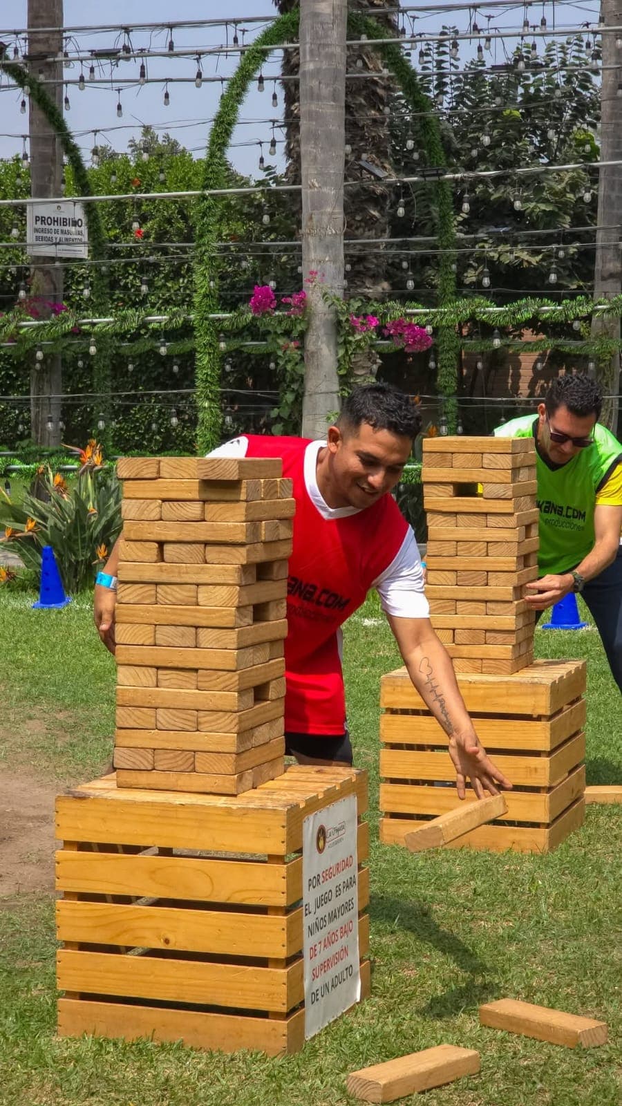 Dos hombres juegan al Jenga gigante sobre cajas de madera en un jardín soleado.