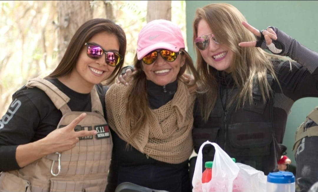 Tres mujeres con gafas de sol y chalecos tácticos sonríen juntas en el exterior.