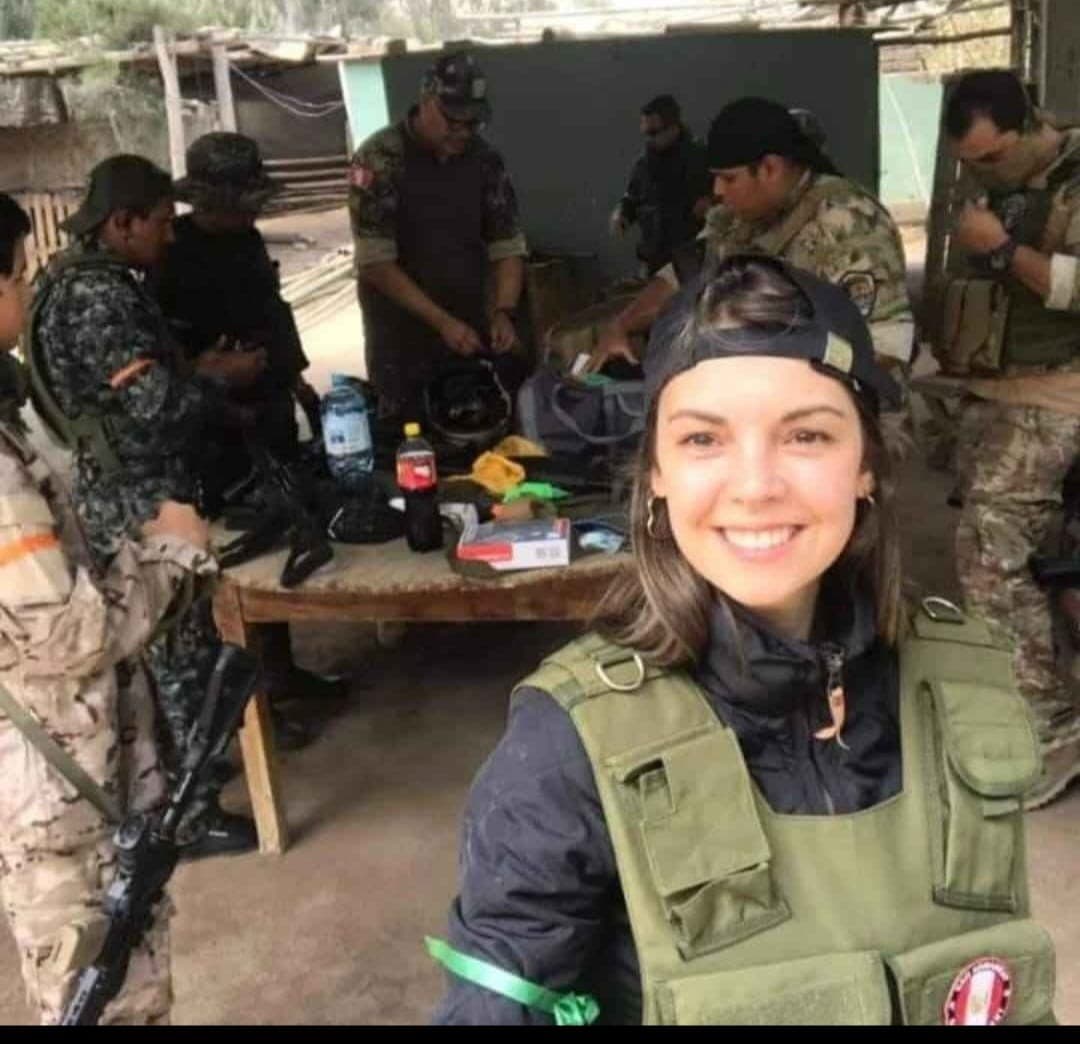 Mujer sonriente con chaleco táctico verde frente a militares que organizan equipo en una mesa.