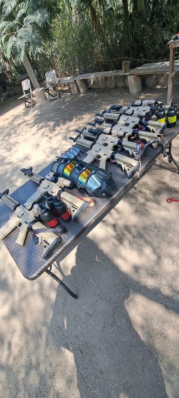 Rifles de airsoft color arena y máscaras tácticas con lentes iridiscentes sobre una mesa exterior.