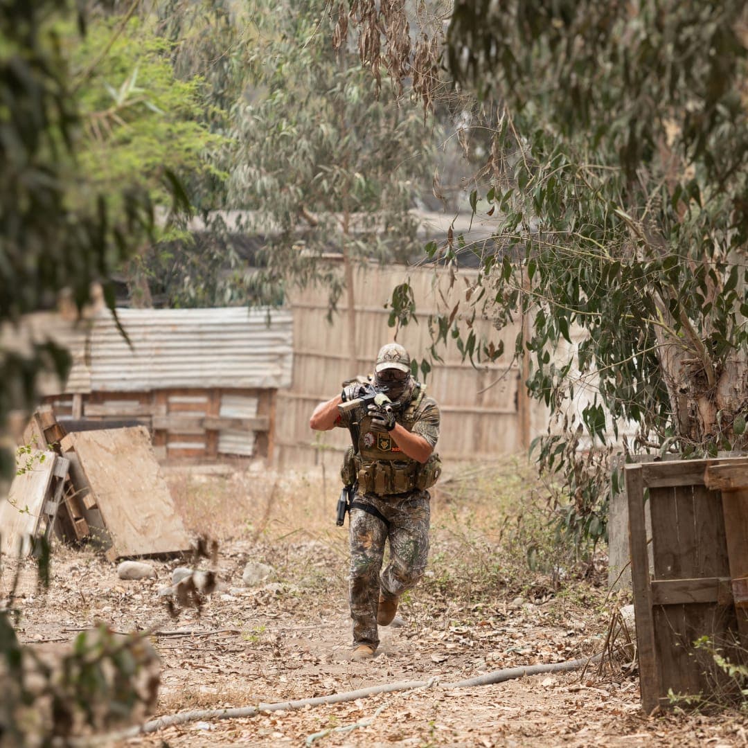 Persona con equipo táctico de camuflaje corriendo y apuntando un rifle en un entorno boscoso.
