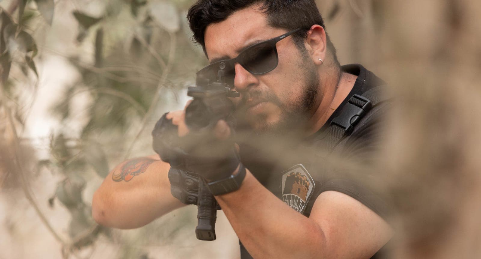 Hombre con gafas de sol apuntando un rifle táctico a través de la vegetación.